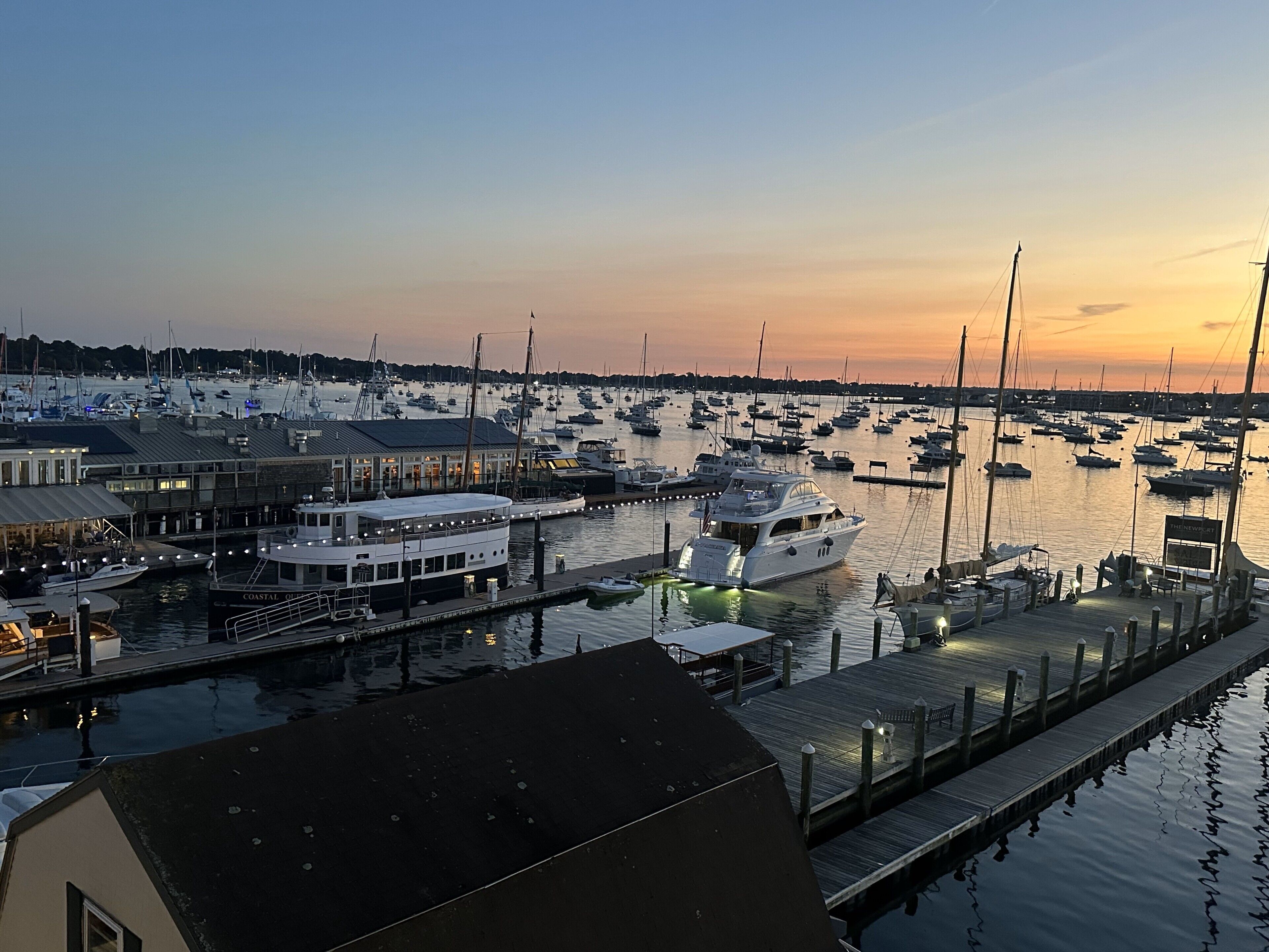 A marina with multiple boats docked at piers, a building with a sign reading "THE NEWPORT," and a wooden walkway along the water.