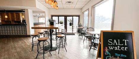 A modern cafe with wooden tables and chairs, a chalkboard sign reading "SALT water," and large windows offering a view of the city.
