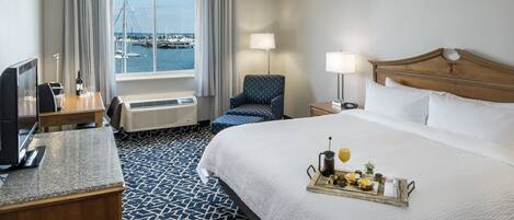 A hotel room with a bed, a desk, a chair, a TV, and a window with a view of the marina.