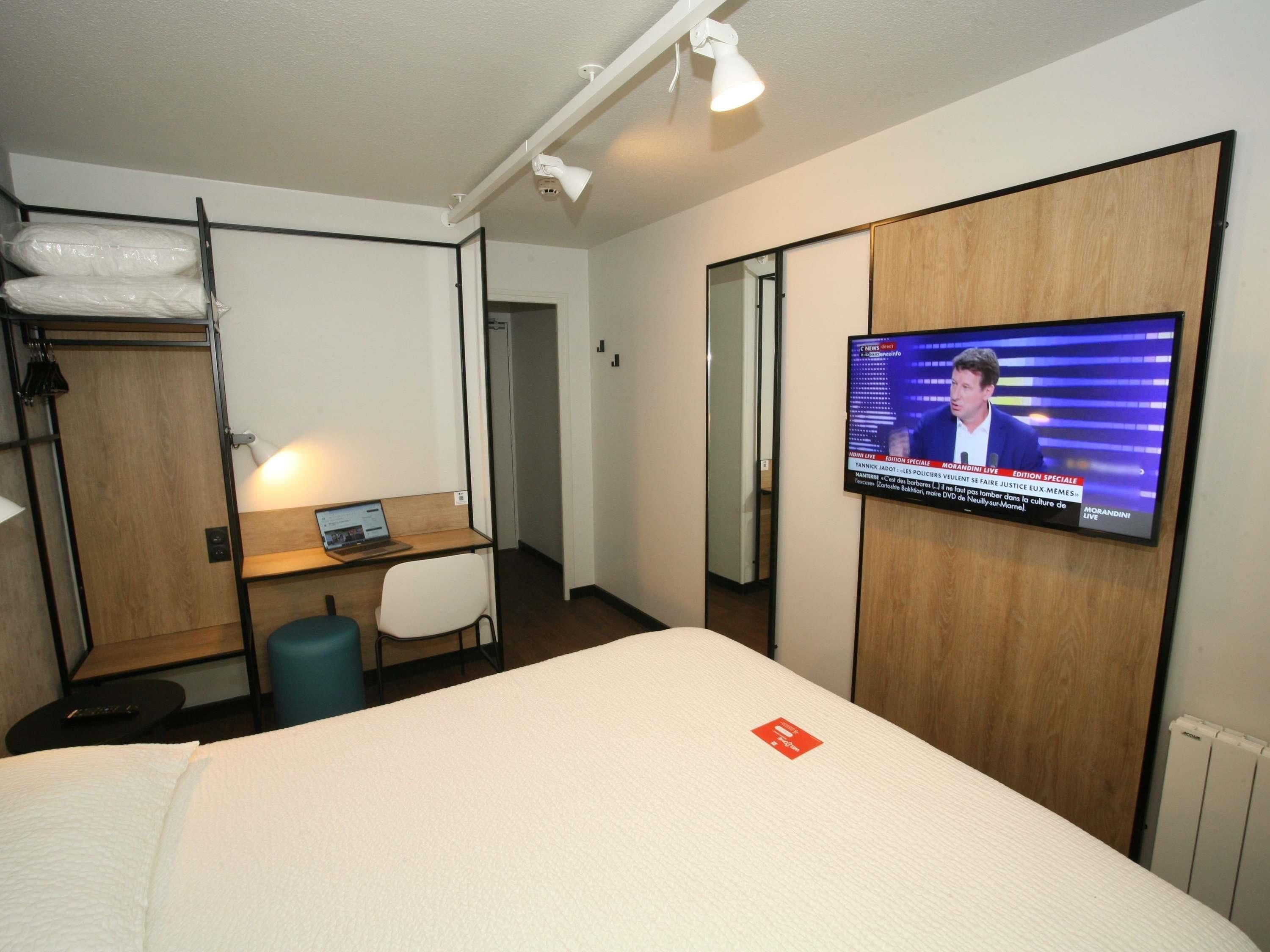 A hotel room with a bed, a desk with a laptop, a TV on the wall, and a wardrobe.