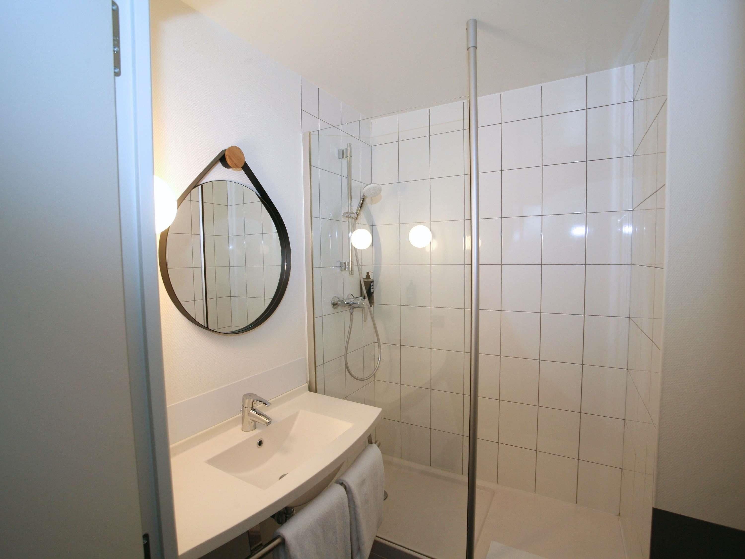 A bathroom with a white sink, a round mirror, a glass shower enclosure, and white tiled walls.