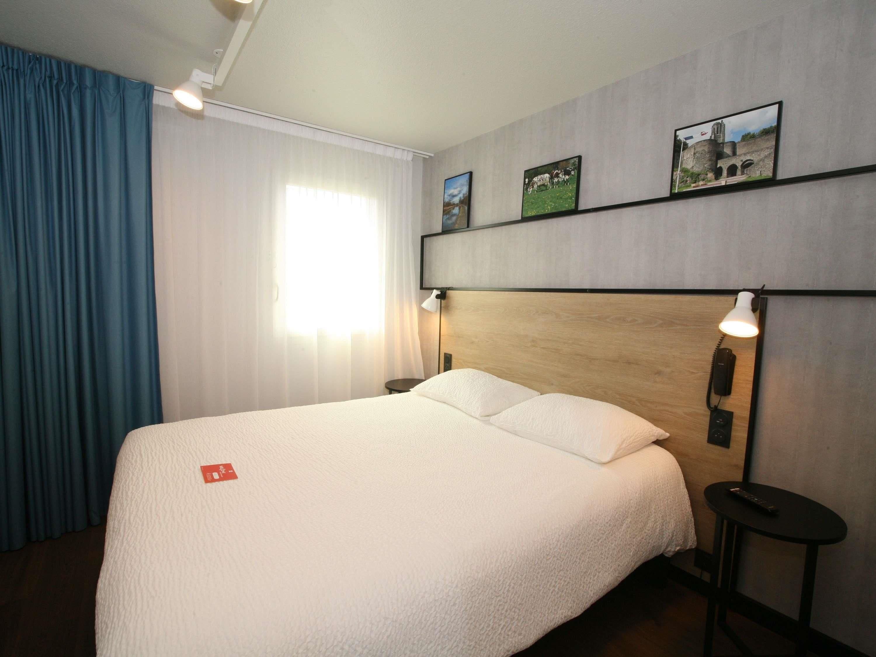 Habitación de hotel con una cama grande, un cabecero de madera, dos lámparas de noche, una mesita pequeña y una ventana con cortinas.