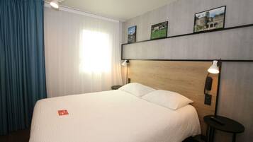Habitación de hotel con una cama grande, un cabecero de madera, dos lámparas de noche, una mesita pequeña y una ventana con cortinas.