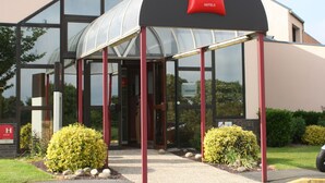 A modern hotel entrance with a glass canopy, red pillars, and a sign for ibis hotels Saint Lô.