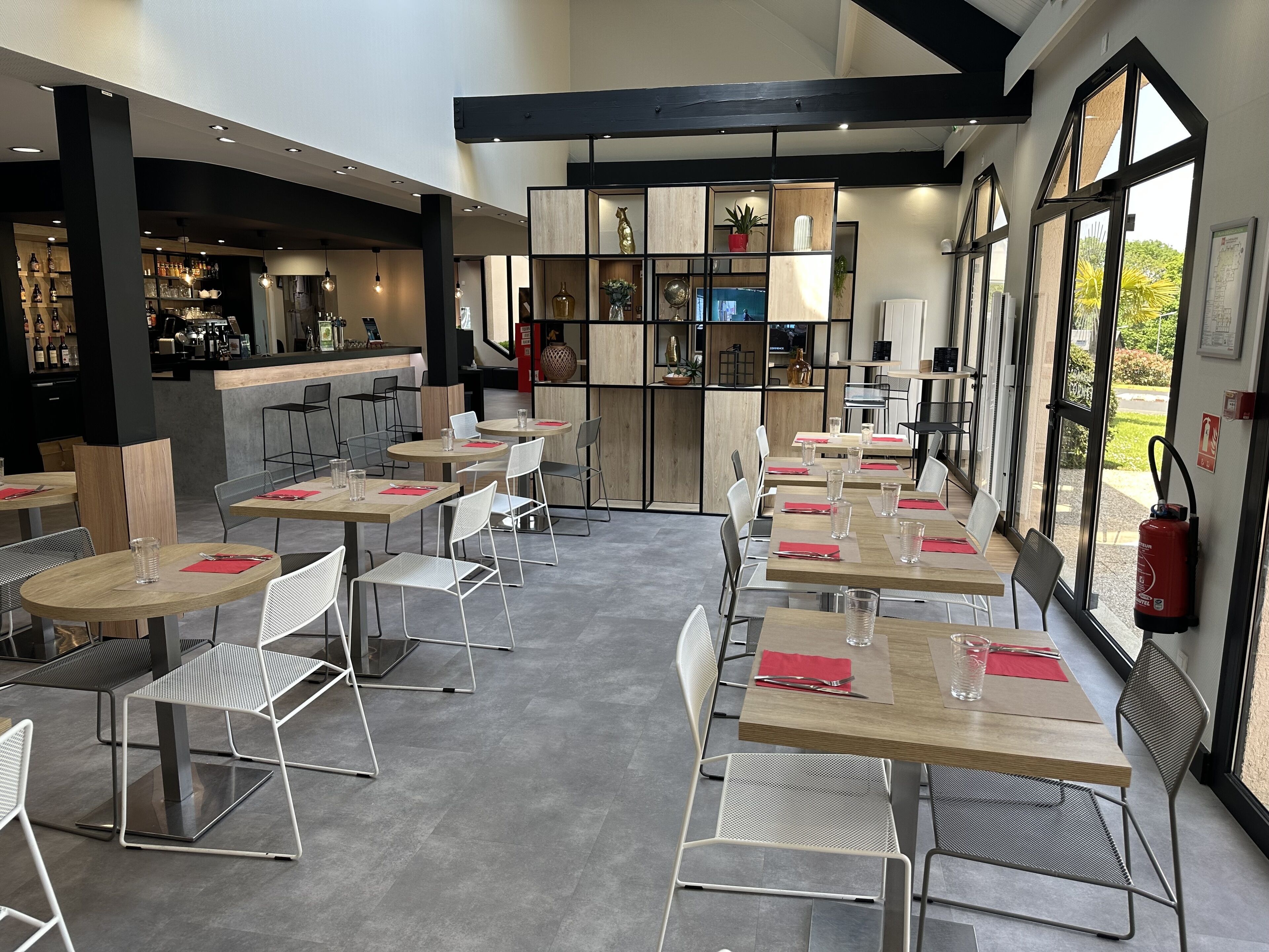 Un restaurante moderno con varias mesas y sillas, una zona de bar y grandes ventanales que ofrecen vistas al exterior.