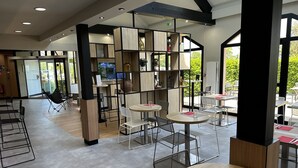 A modern indoor dining area with round tables and metal chairs, wooden and black metal shelving units, and a view of greenery outside.