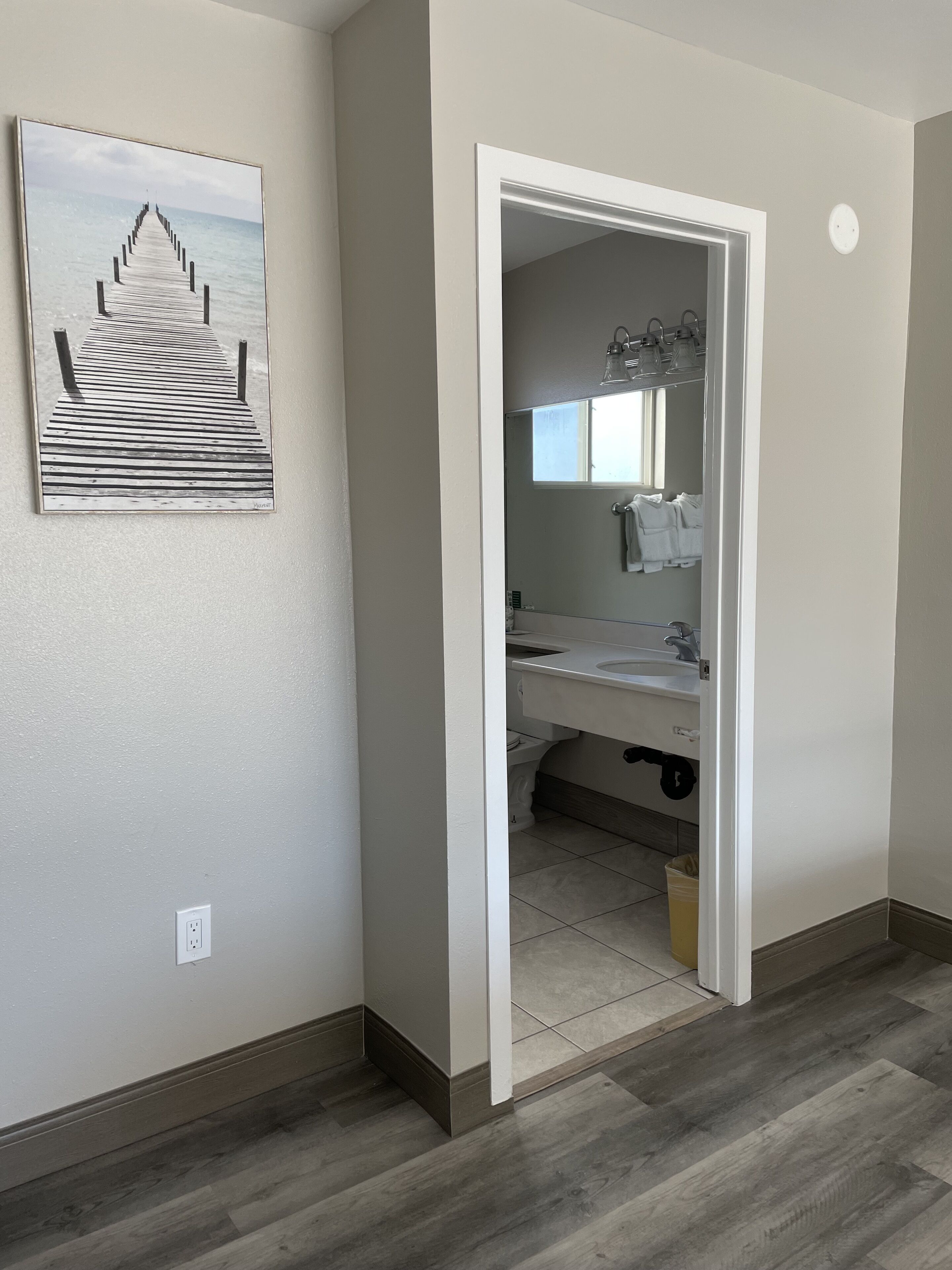 A hallway with a wooden floor, a door leading to a bathroom with a sink and mirror, and a framed picture on the wall.