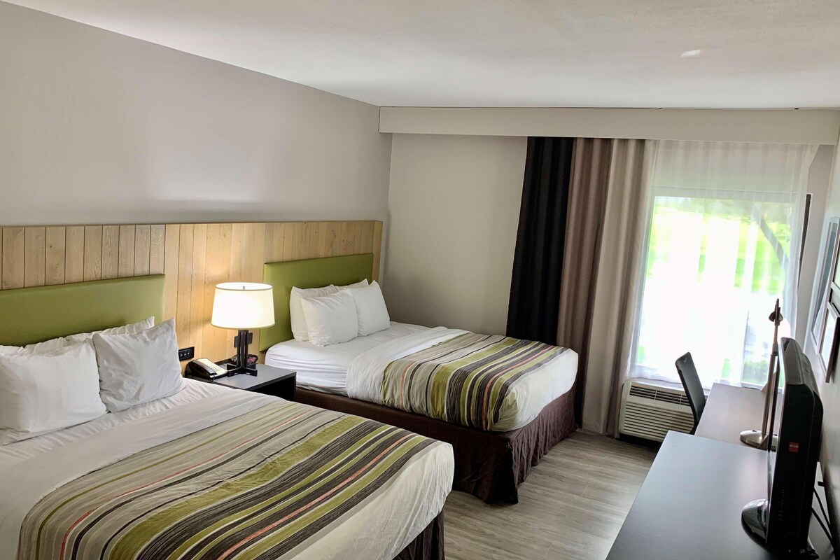 A hotel room with two beds, a desk with a computer, a lamp, and a window with curtains.