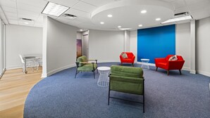 Una sala de reuniones moderna con alfombra azul, paredes blancas y una pared de acento en azul. Hay dos sillones verdes, dos sillones rojos y una mesa blanca con estructura de alambre.