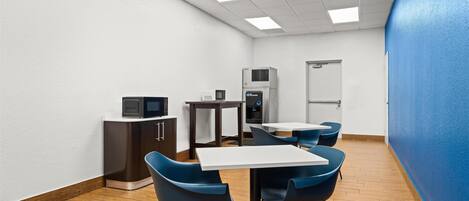 A modern, well-lit room with a white ceiling, wooden flooring, and blue chairs around a central table.