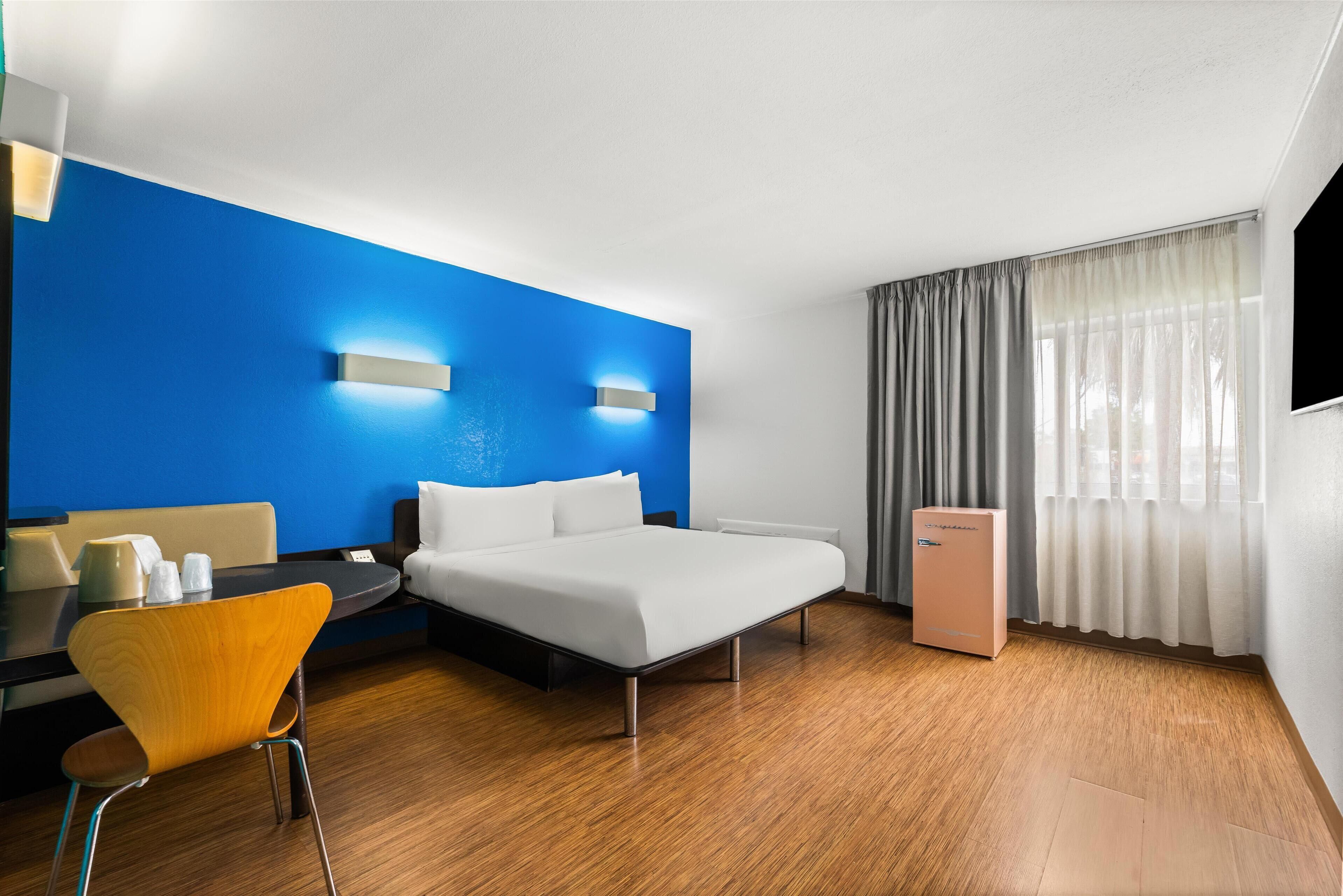 Une chambre d’hôtel moderne avec un plancher en bois, un lit, un bureau avec une chaise, un petit réfrigérateur et une fenêtre avec des rideaux.