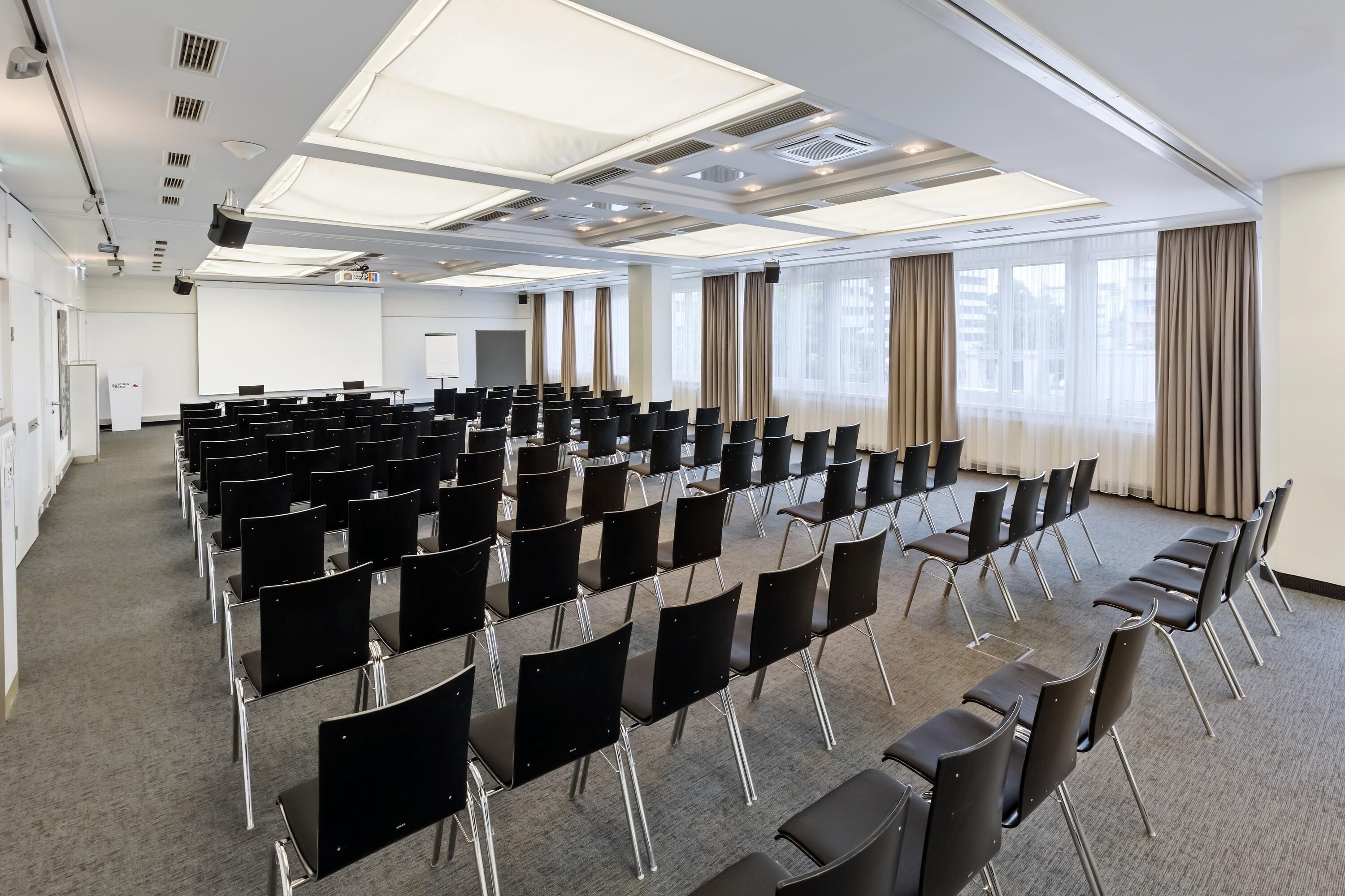 A conference room with rows of black chairs, a projector screen, and large windows with curtains.