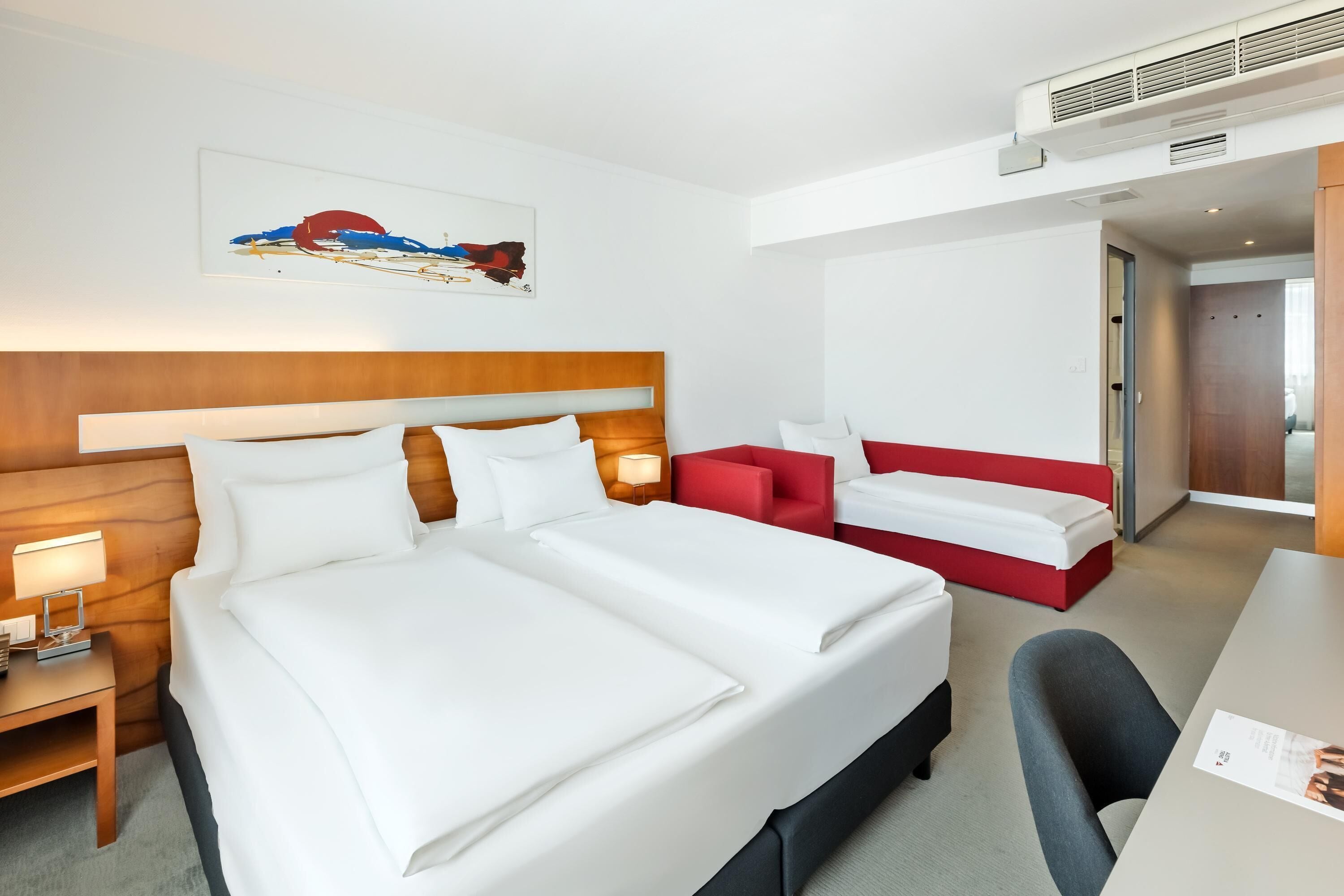 A hotel room with a large bed, a desk, a chair, a red sofa, and a painting on the wall.