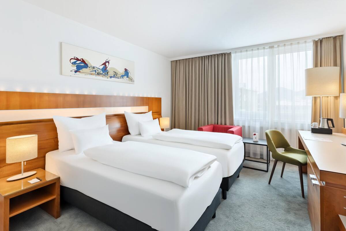 A hotel room with two beds, a desk, a chair, a lamp, and a painting on the wall.