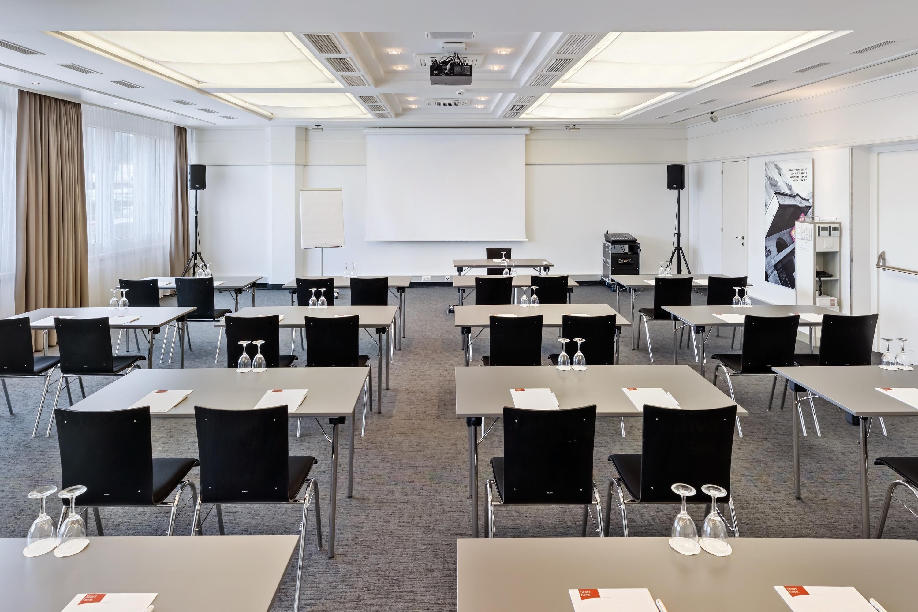 Un salón de conferencias con varias mesas y sillas, una pantalla de proyector y una pizarra.