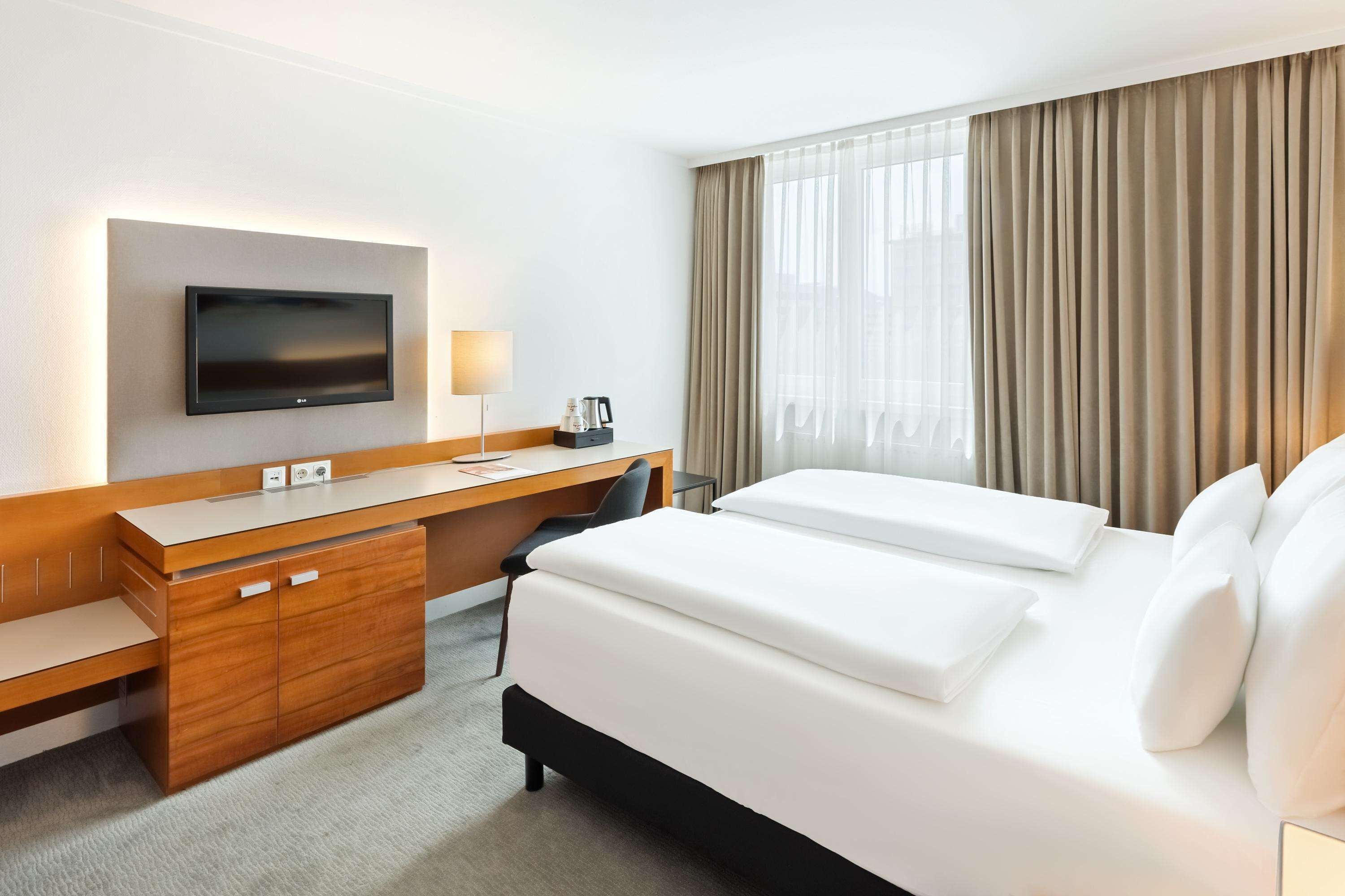 A hotel room with a bed, a desk, a chair, a TV, and a window with curtains.