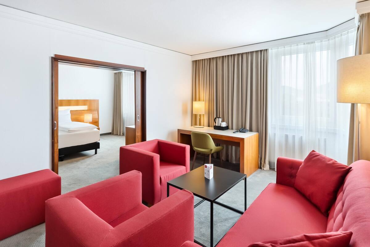 A hotel room with a red sofa, a desk with a lamp, a bed, and a large window with curtains.