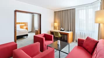 Habitación de hotel con sofá rojo, un escritorio con lámpara, cama y un ventanal con cortinas.