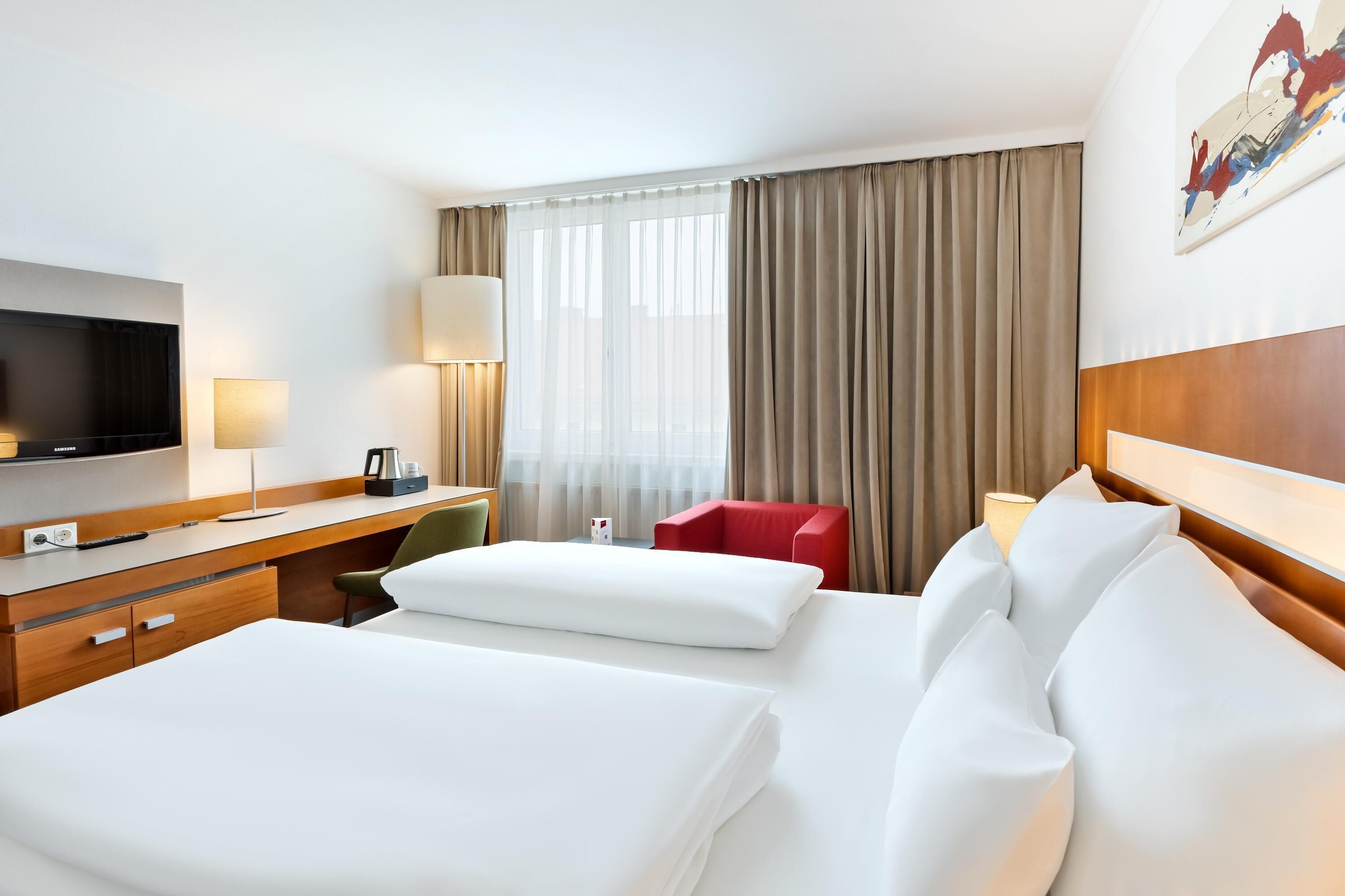 Quarto de hotel com uma cama grande, uma mesa de trabalho com televisão, uma cadeira vermelha e uma janela ampla com cortinas.