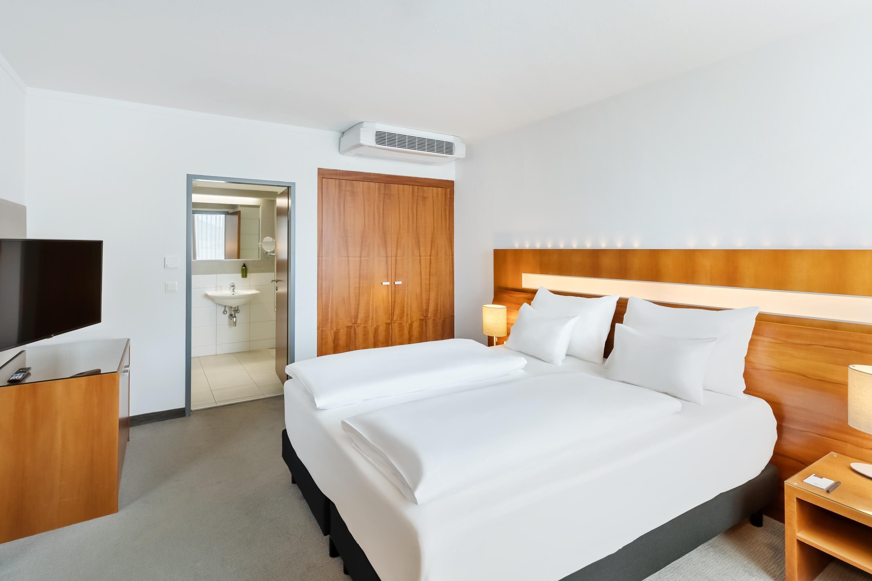 Ein Hotelzimmer mit einem großen Bett, einem Holzkopfteil, einem Nachttisch mit Lampe, einem Fernseher auf einem Schreibtisch und einem durch eine offene Tür einsehbaren Badezimmer.