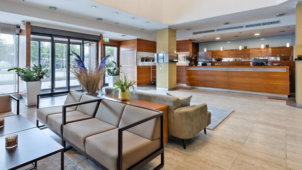 A modern hotel lobby with a reception desk, seating area, and a kitchen in the background. - Austria Trend Hotel Europa Graz Hauptbahnhof (Graz)