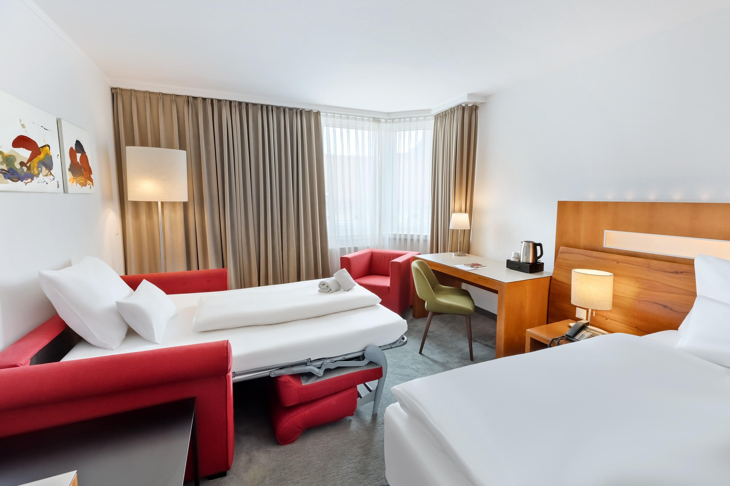 Una habitación de hotel con cama, escritorio, silla, sofá y ventana con cortinas.