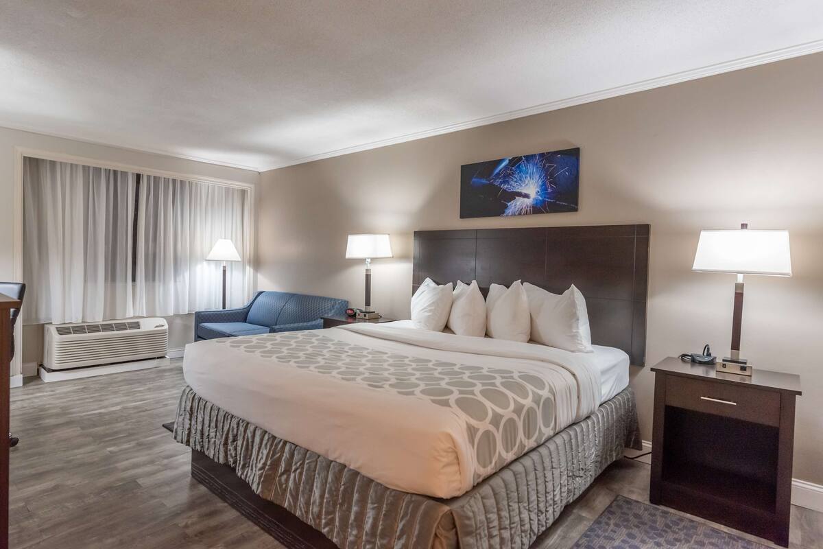 Une chambre d’hôtel avec un grand lit, deux tables de chevet avec des lampes, une chaise bleue, une grande fenêtre avec des rideaux et une œuvre d’art accrochée au mur.