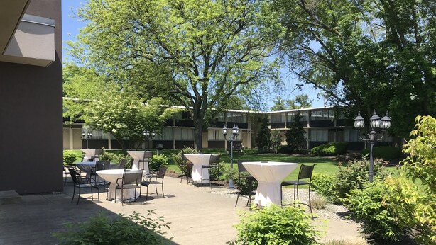 Een terras buiten met tafels en stoelen, omringd door groen en een gebouw op de achtergrond.
