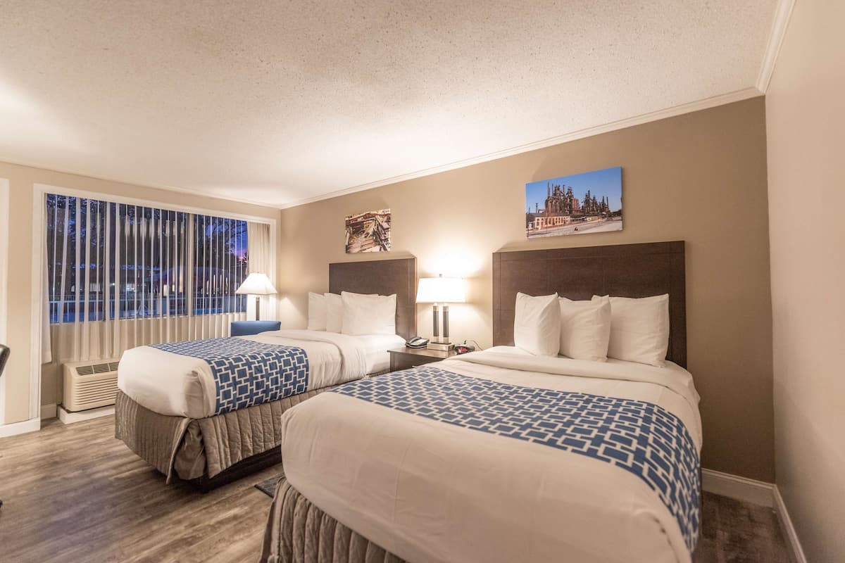 Une chambre d’hôtel avec deux lits, une grande fenêtre avec stores, une lampe fixée au mur et un tableau encadré.