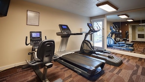 Een hotel fitnessruimte met een loopband, elliptische trainer en hometrainer, plus een scherm en een spiegel die de apparatuur weerspiegelt.