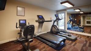 Een hotel fitnessruimte met een loopband, elliptische trainer en hometrainer, plus een scherm en een spiegel die de apparatuur weerspiegelt.