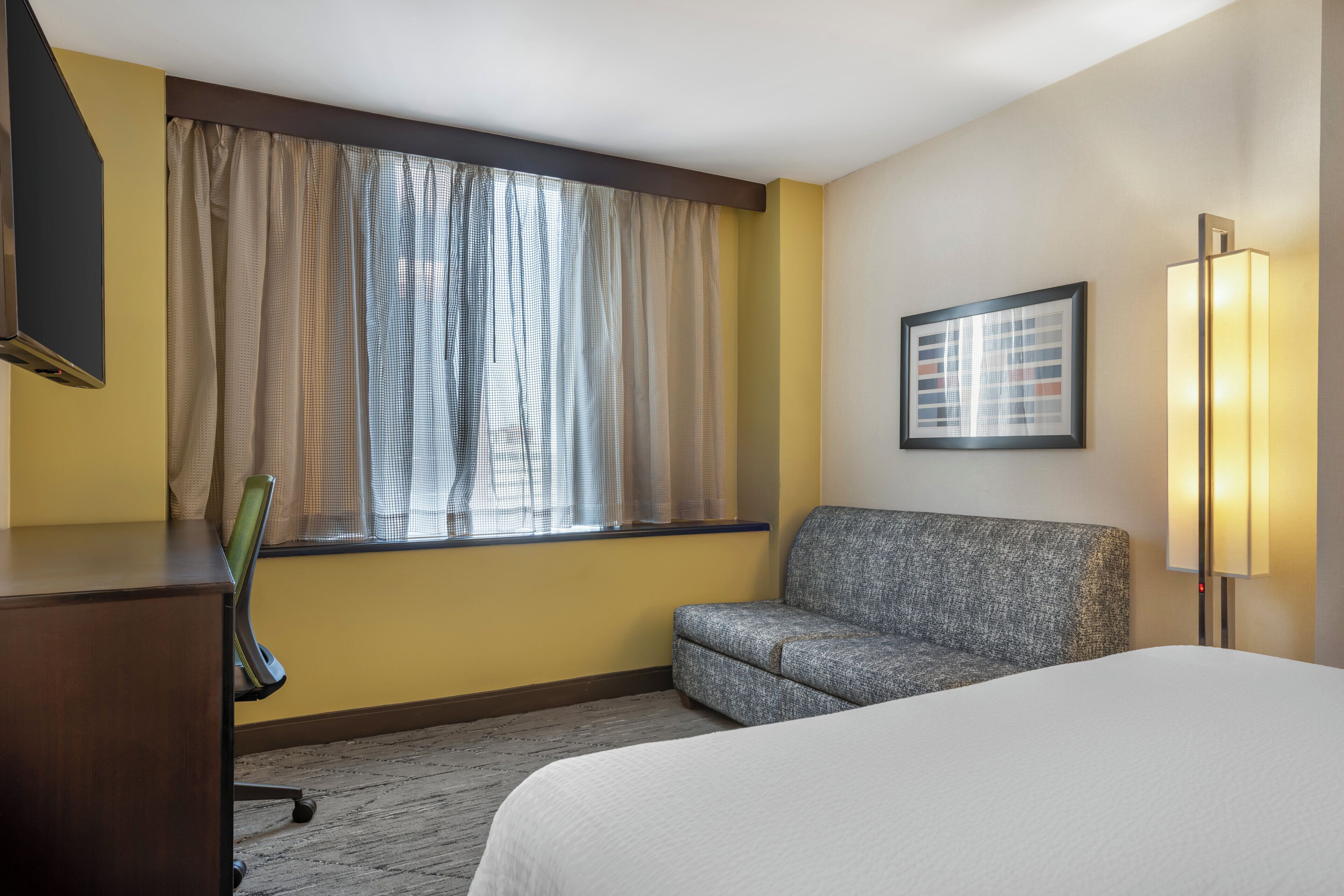 Habitación de hotel con cama, sofá, escritorio, silla, ventana con cortinas y un cuadro en la pared.