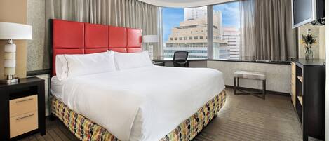 A hotel room with a large bed, a desk, a chair, a TV, and a view of the cityscape.