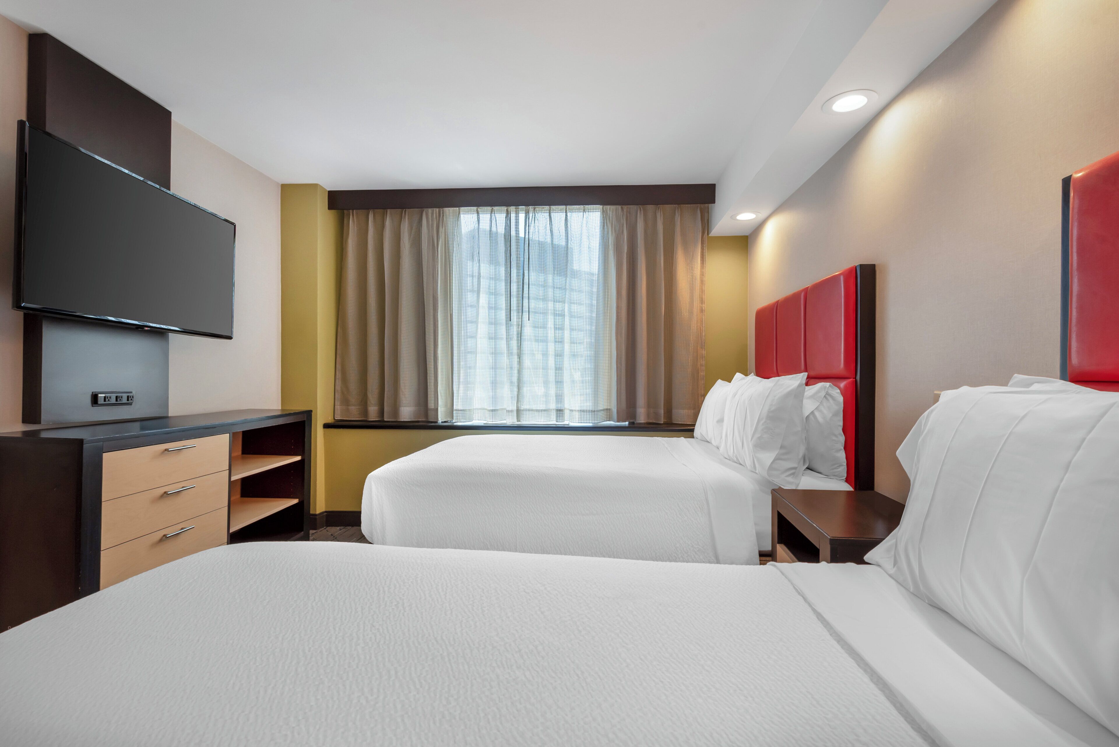 A hotel room with two beds, a flat-screen TV, a desk, and a window with curtains.