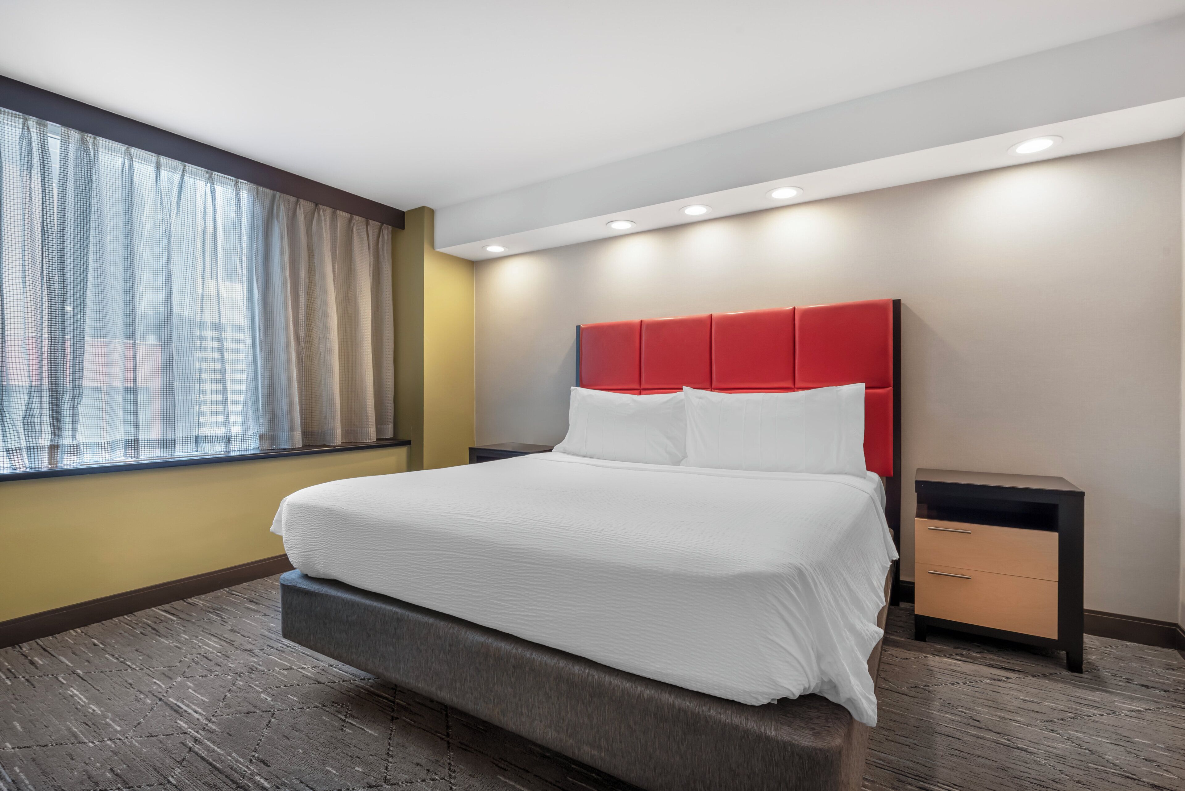 A hotel room with a large bed, a nightstand, a window with curtains, and a modern ceiling design.