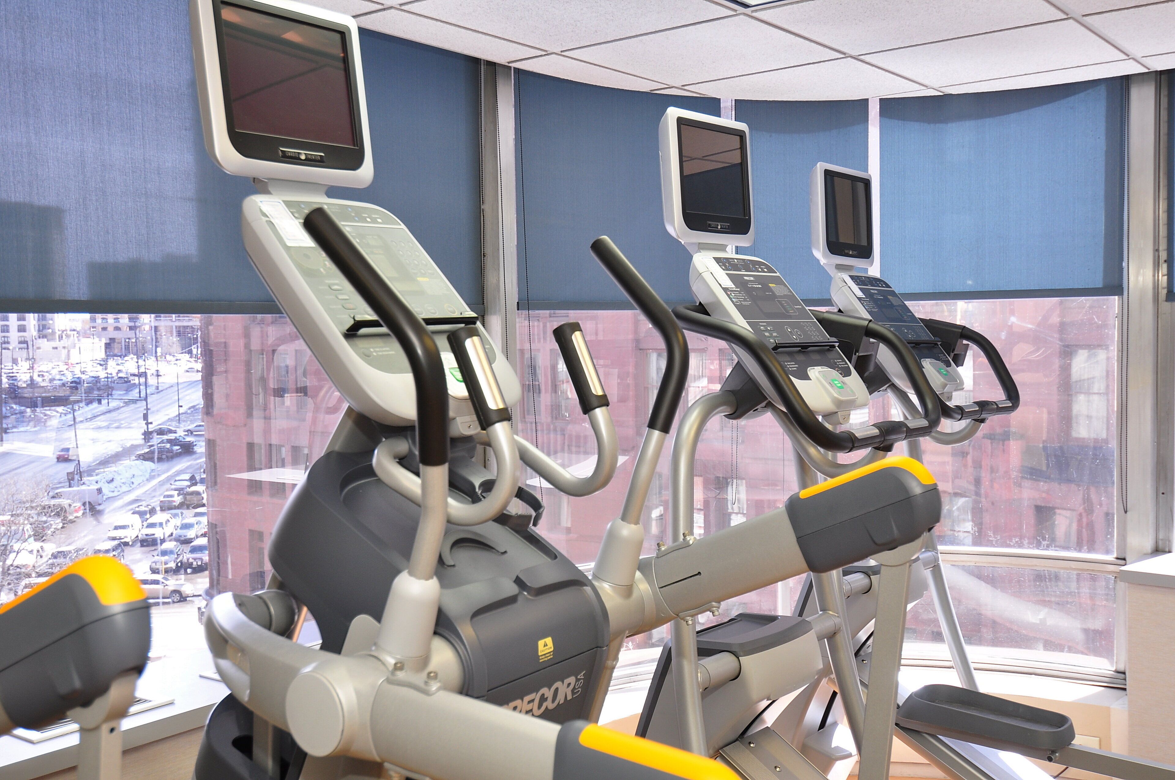 Ett gym med löpband och annan träningsutrustning, stora fönster med utsikt över staden och en blå vägg.