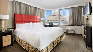 A hotel room with a large bed, a desk, a chair, a TV, and a view of the city.