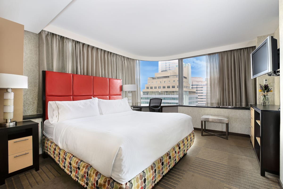 A hotel room with a large bed, a desk, a chair, a TV, and a view of the city.