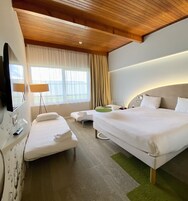 Habitación de hotel con una cama grande, una cama más pequeña, un escritorio con espejo, un televisor y una ventana con cortinas.