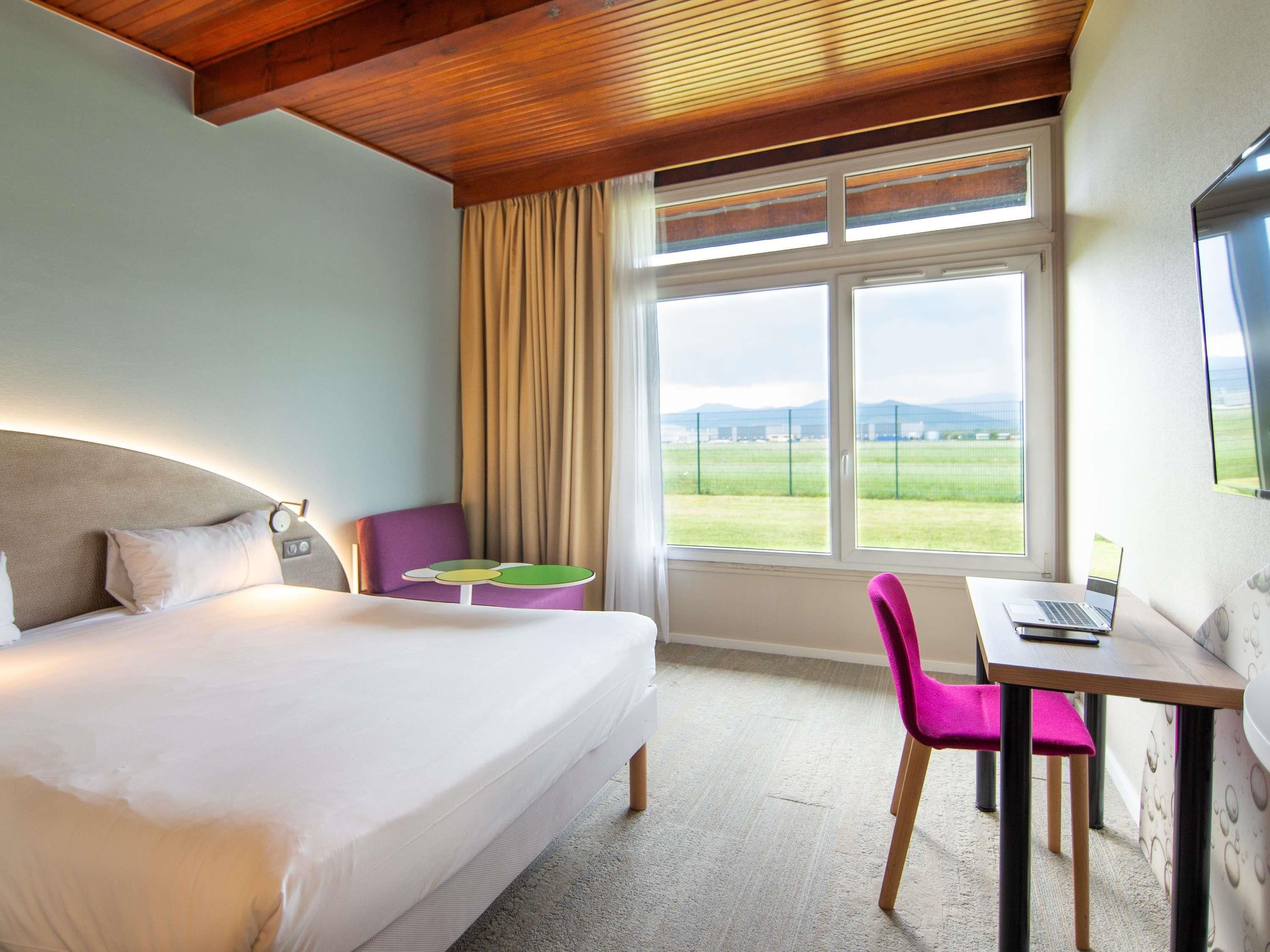 Une chambre d’hôtel avec un lit, un bureau avec un ordinateur portable, une chaise et une fenêtre donnant sur la campagne.
