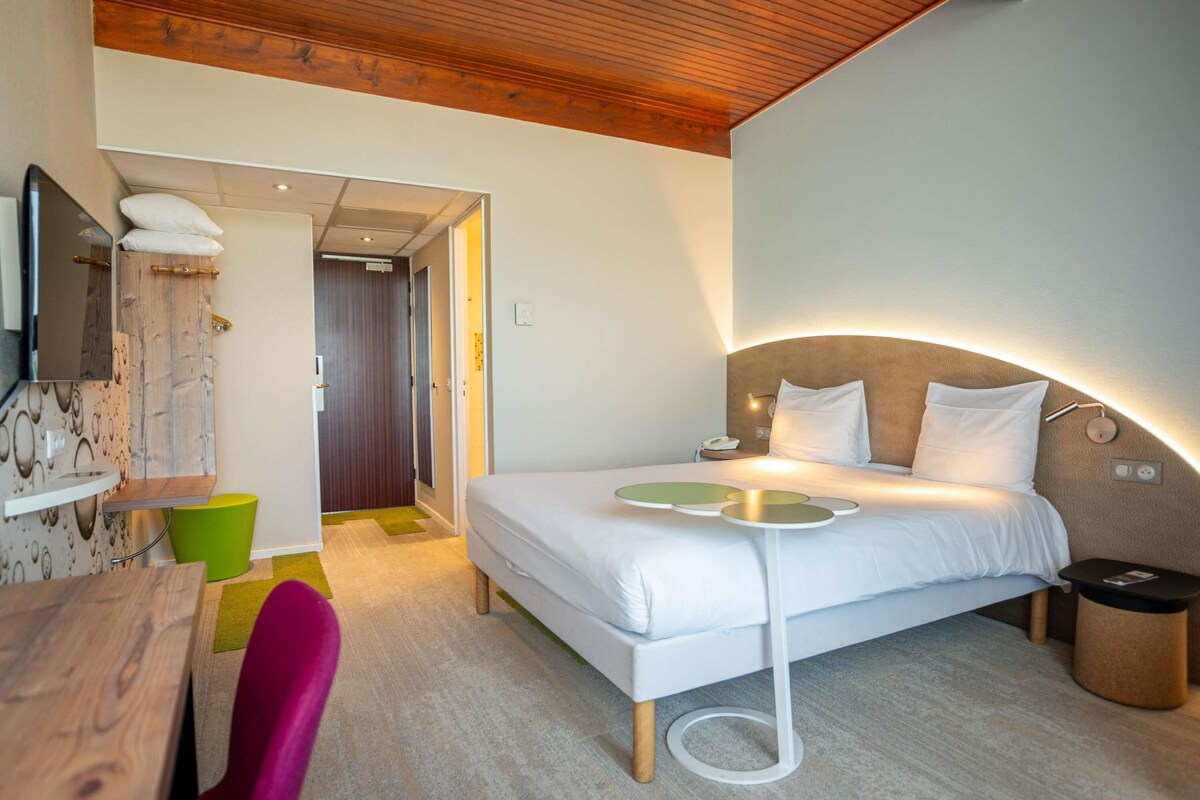 A modern hotel room with a bed, bedside table, chair, and a TV mounted on the wall.