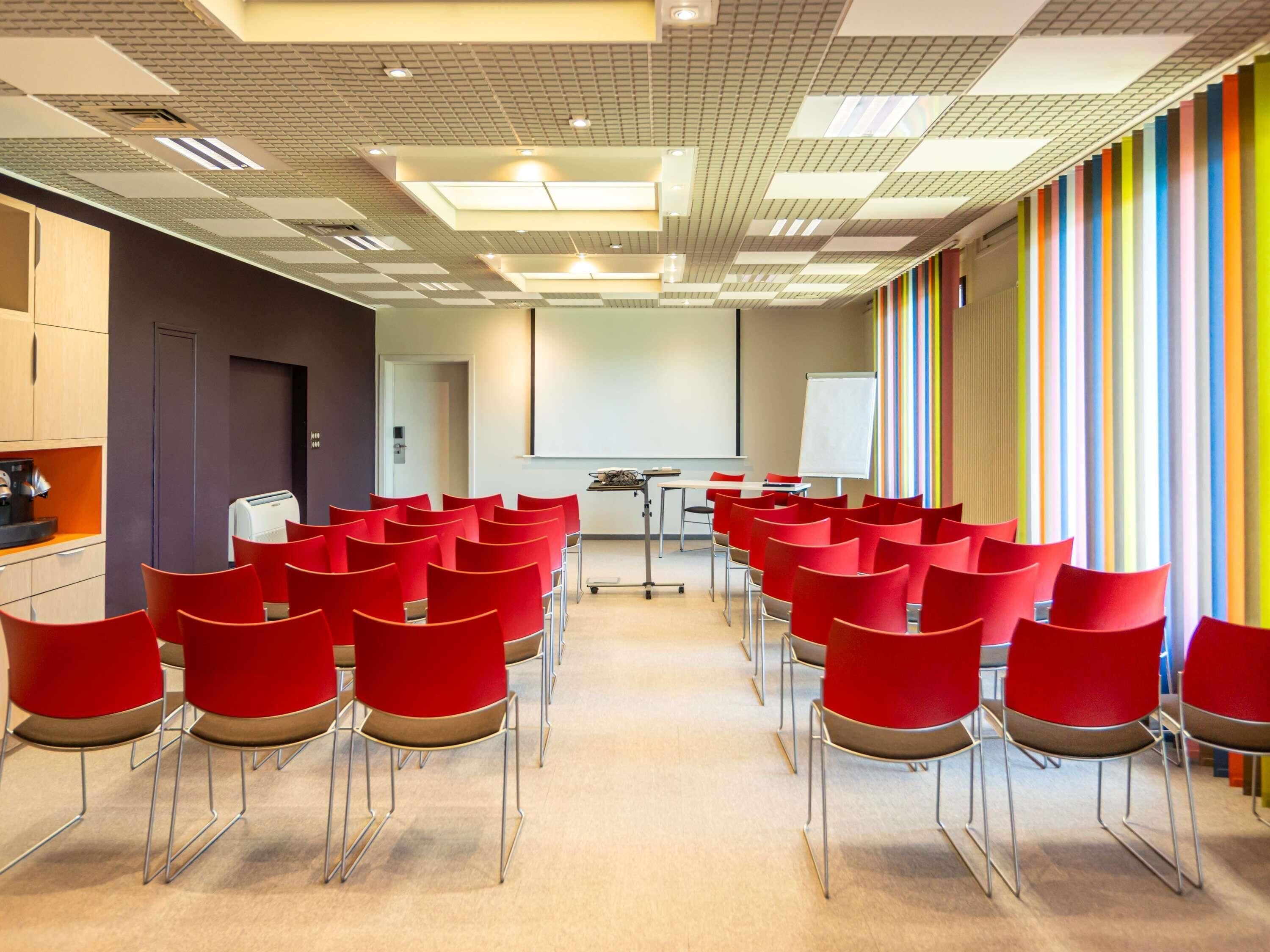 Une salle de conférence avec des chaises rouges, un tableau blanc et un écran de projection.