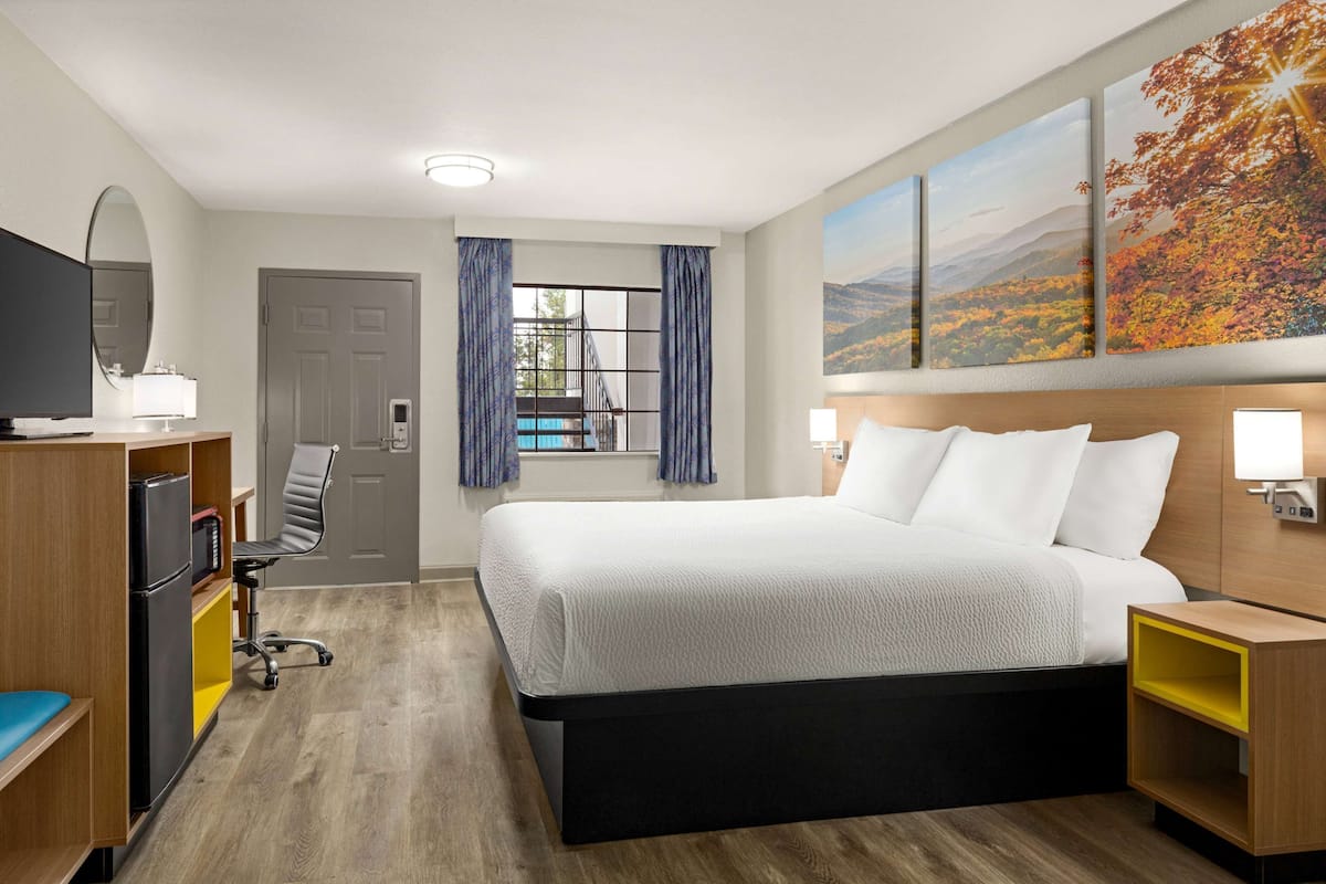 A hotel room with a bed, desk, chair, TV, and artwork on the wall.