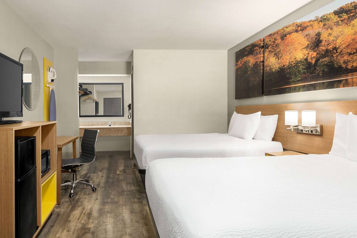 A hotel room with two beds, a desk with a chair, a refrigerator, a microwave, a mirror, and a painting of a forest.