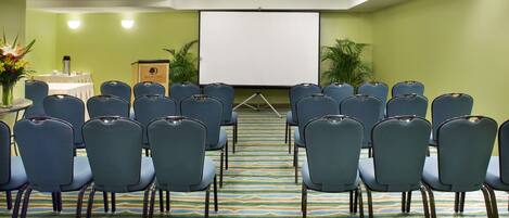 Un salón de conferencias con filas de sillas azules, una pantalla de proyector y un atril.