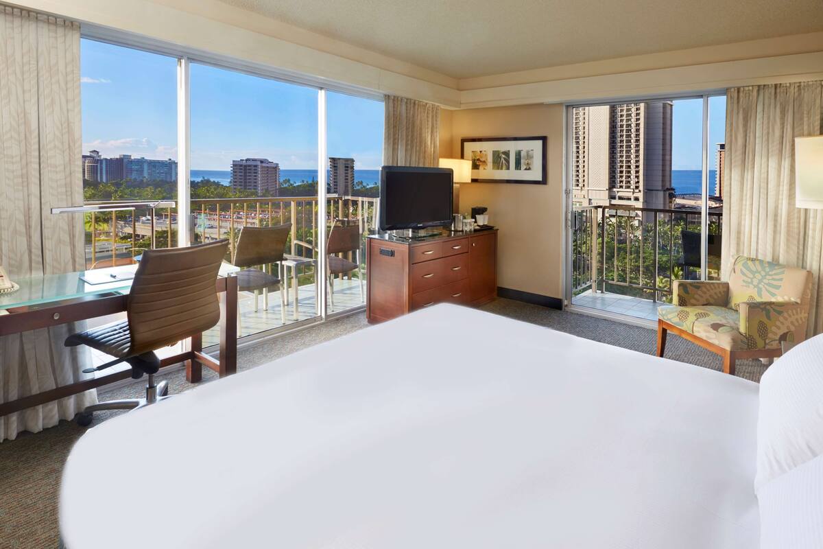 A hotel room with a large bed, a desk with a chair, a television, and a balcony with a view of the city and ocean.