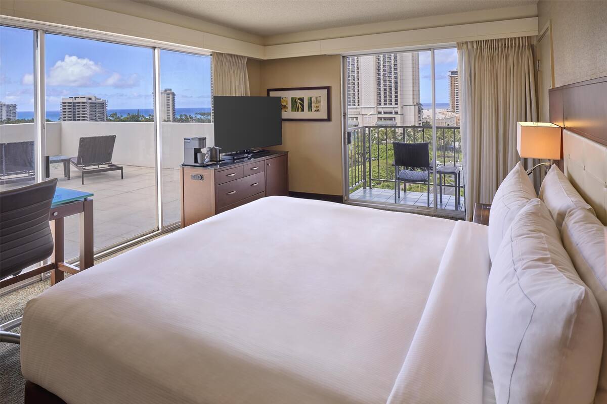 A hotel room with a large bed, a desk with a chair, a TV, and a balcony with a view of the city and ocean.