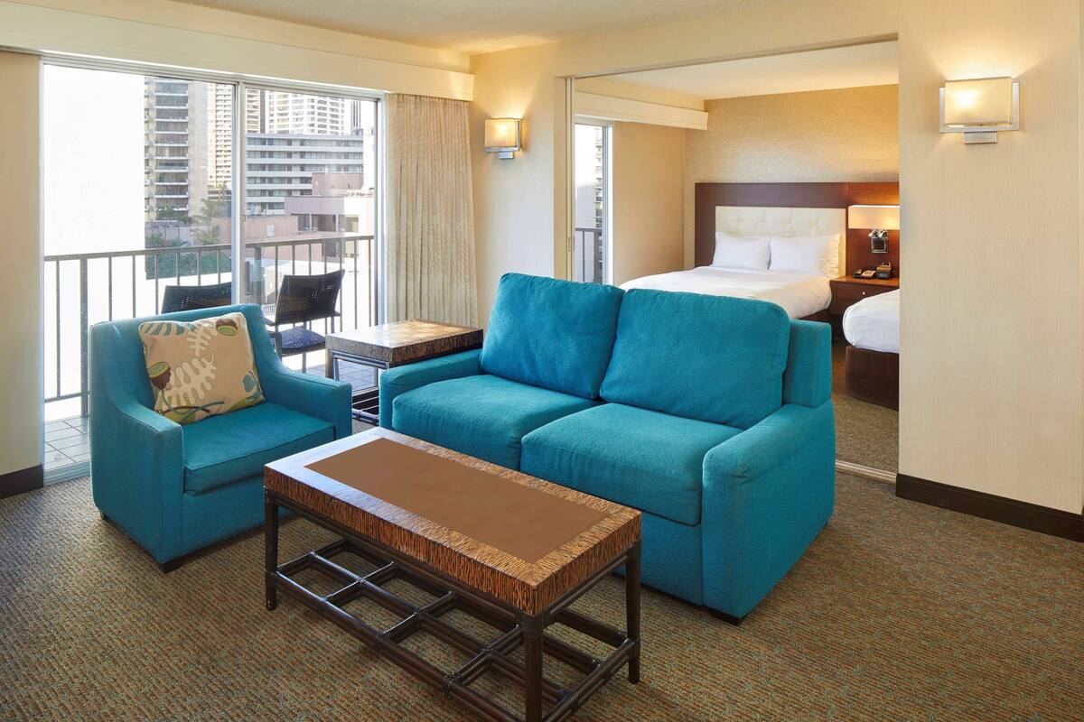 A hotel room with a large bed, a blue sofa, a small table, and a balcony with a view of the city.
