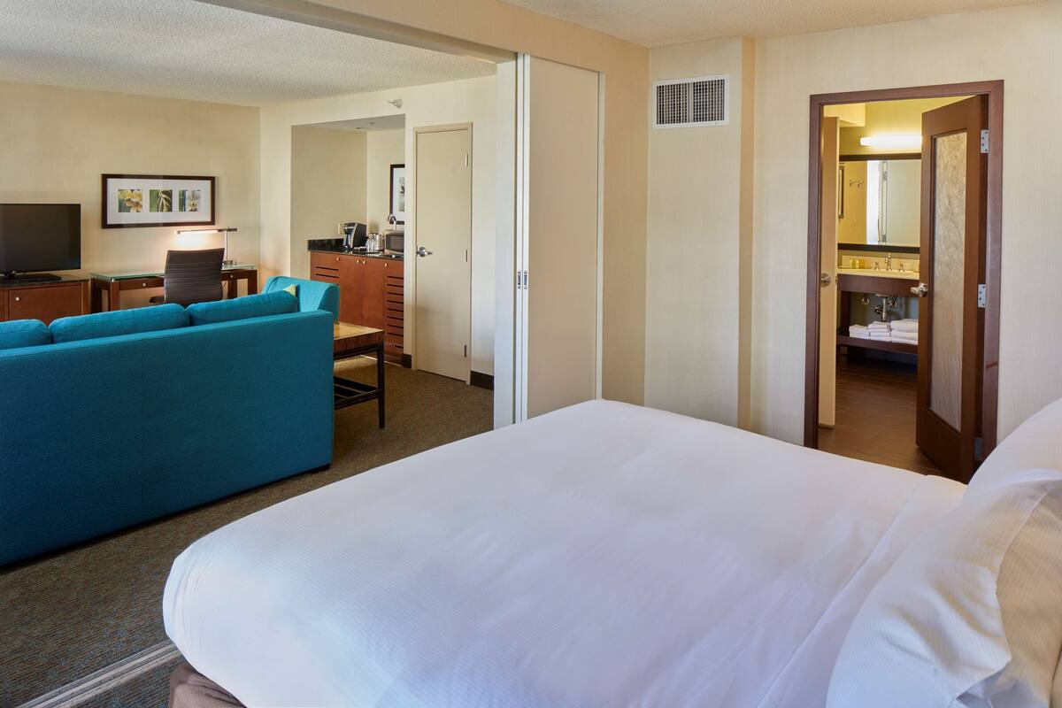 A hotel room with a large bed, a blue sofa, a desk with a chair, a television, and a bathroom visible through an open door.
