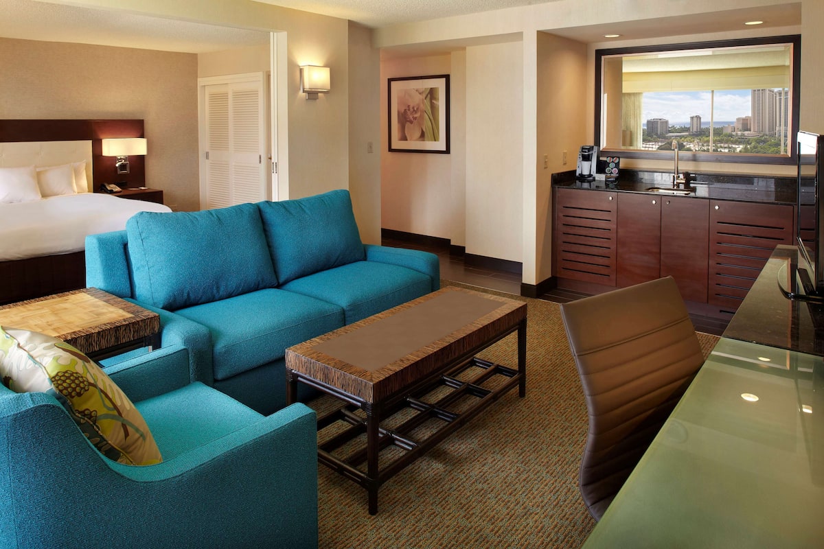 A hotel room with a bed, a blue sofa, a wooden coffee table, a glass dining table, and a view of the city through a window.